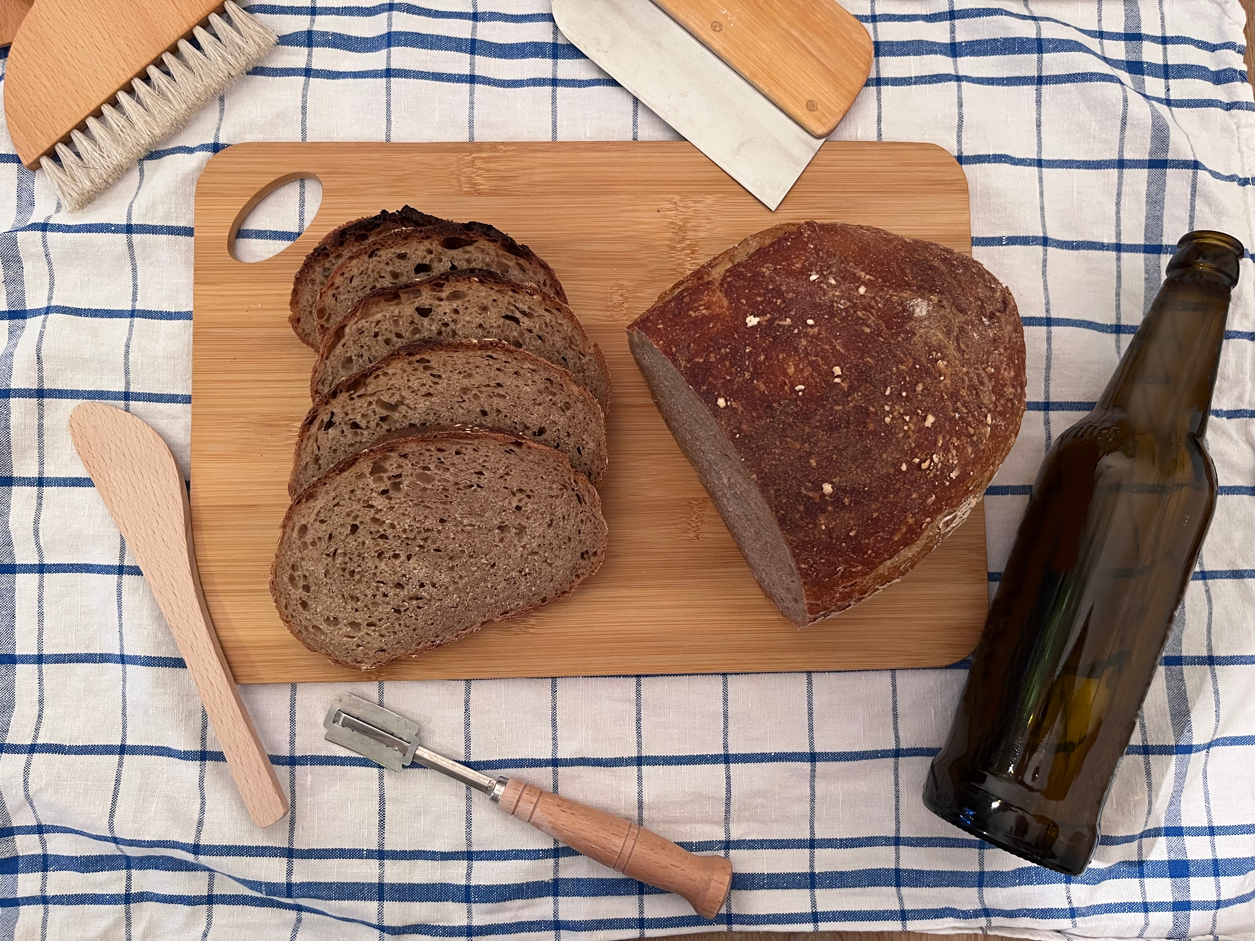 A dark black beer bread with a porous crumb lies on a wooden board, next to baking utensils and a beer bottle