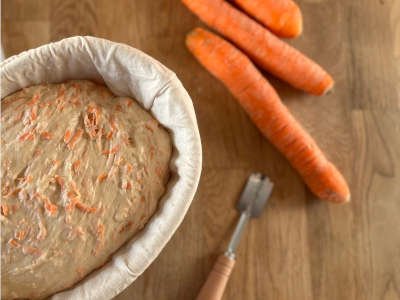 A carrot bread dough lies in a proofing basket, next to it are 3 carrots and a dough knife