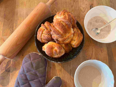 A bowl of delicious cinnamon vanilla knots stands on a wooden table, next to it love various baking utensils