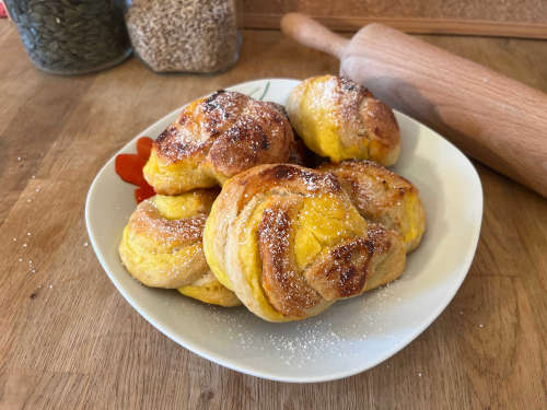 Gistbroodjes met vlaaivulling gestapeld op een bord en bestrooid met poedersuiker. Op de achtergrond staan een deegroller en twee potten met bakingrediënten.