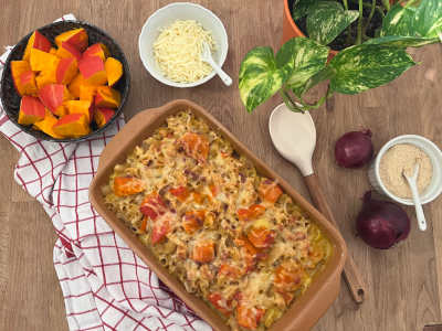 Auf einem Holztisch steht eine Auflaufform mit einem überbackenen Kürbis-Pasta-Auflauf, daneben stehen Schüsseln mit Streukäse und Hefeflocken