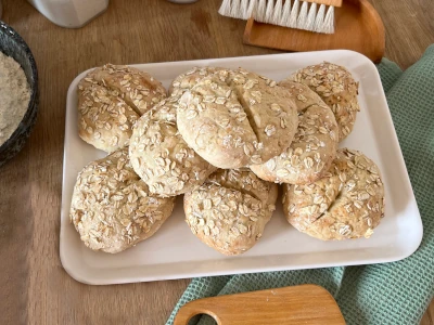 A tray with 9 wheat rolls with oatmeal decoration lies on a wooden table, next to it a green kitchen towel and various baking utensils