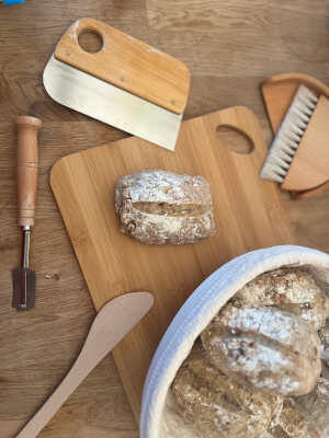 Een tarwe-spelt-roggebroodje ligt op een houten plank, ernaast ligt wat bakgerei en een mand met meer broodjes