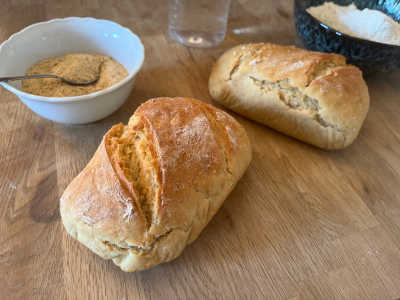 Two mini wheat loaves lie on a wooden table, in the background a small bowl with yeast flakes and a larger bowl with wheat flour