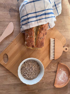 A spelt bread decorated with some sunflower seeds lies on the wooden board, next to it a bowl with sunflower seeds and other baking utensils