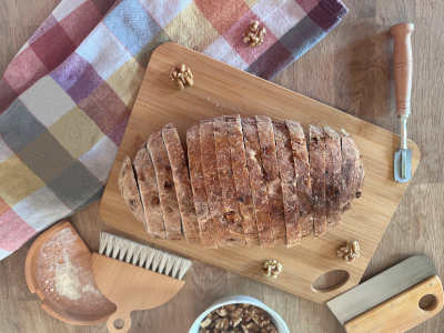 Una rebanada de pan de espelta con nueces reposa sobre una tabla de madera, junto a ella algunas nueces y otros utensilios de panadería