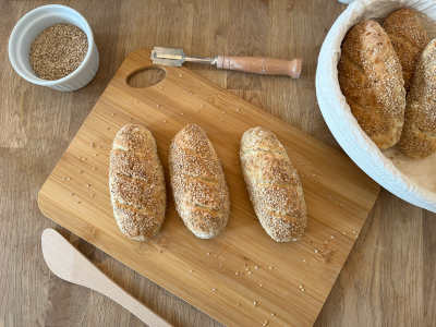 Spelt- en sesamstengels op een houten plank, daarnaast een broodmandje met meer broodjes en een schaaltje sesamzaadjes