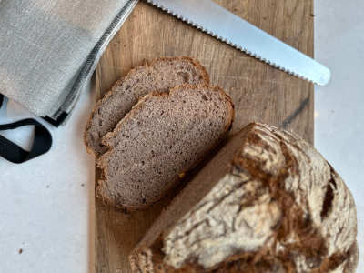 Un pane scuro di farro e noci con una crosta di farina giace su una tavola di legno, con le prime due fette tagliate. Accanto al pane ci sono un coltello da pane e un sacchetto per il pane.