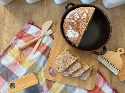 En halv speltlimpa ligger i en brödform, bredvid ligger några skivor spelt- och morotsbröd med linfrö. Runtomkring brödet finns bakredskap och en kökshandduk.