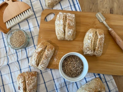 Two-seed rolls are on a wooden board, next to bowls with sesame and sunflower seeds, as well as other baking utensils.