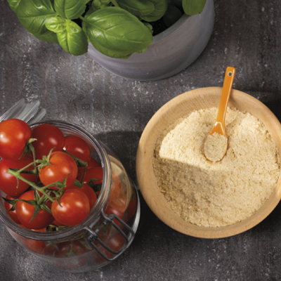 A bowl of vegan parmesan made from yeast flakes and almonds stands on a table, next to it a bowl of tomatoes and a pot of basil.